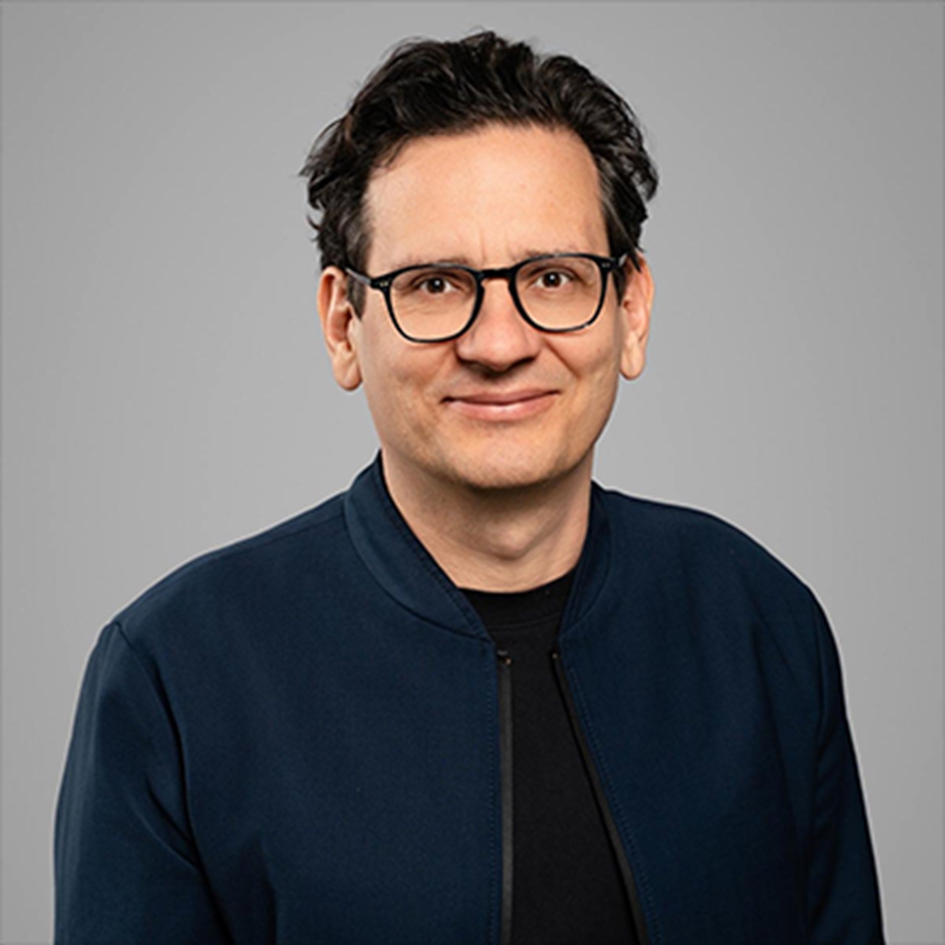 Lächelnder Mann mit schwarzen Haaren und schwarzer Brille, der ein schwarzes T-Shirt und eine dunkelblaue Jacke trägt.