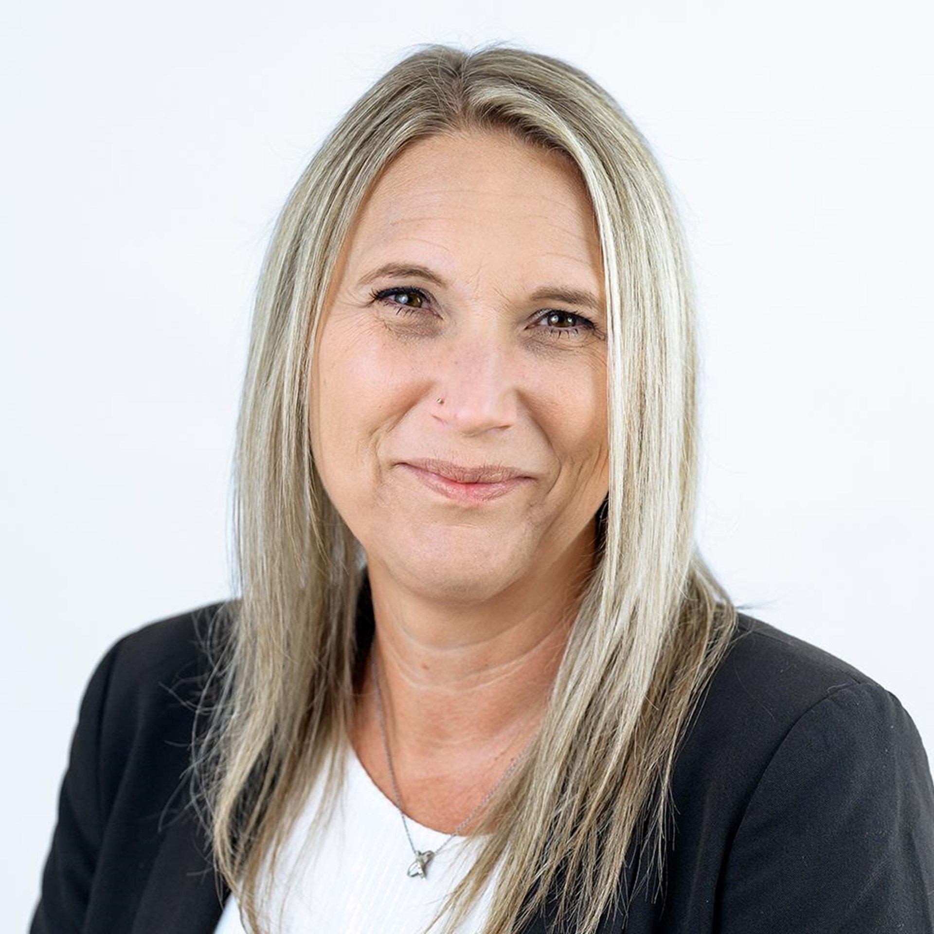 Eine Frau mit glatten, schulterlangen, blondierten Haaren lächelt dezent. Sie trägt einen schwarzen Blazer über einem weißen Oberteil und eine feine Halskette mit kleinem Anhänger vor einem weißen Hintergrund.