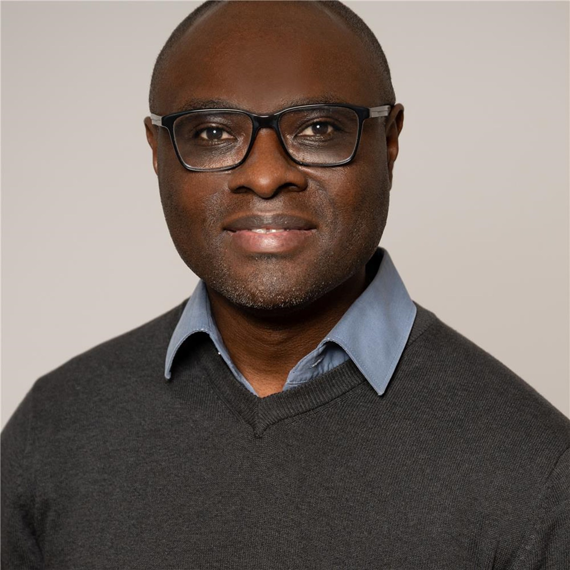 Oberkörperporträt einer Person mit dunkelgrauem Pullover über hellblauem Hemd vor einfarbigem Hintergrund.