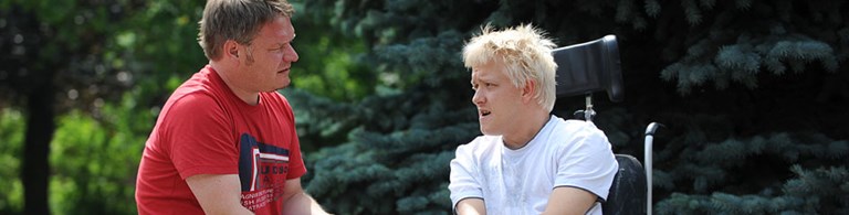 Ein Mann im roten T-Shirt spricht mit einem jungen Mann im Rollstuhl, der blonde Haare hat. Im Hintergrund sind grüne Bäume zu sehen.